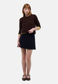 Pull marron avec des finitions jaunes, associé à une jupe bleu marine. Les chaussures présentent un design tissé avec un accent de boucle. Fond blanc uni.