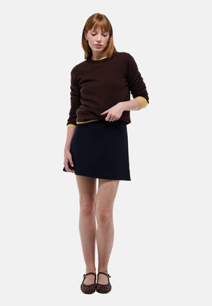 Pull marron avec des finitions jaunes, associé à une jupe bleu marine. Les chaussures présentent un design tissé avec un accent de boucle. Fond blanc uni.