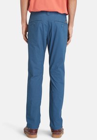 Blauwe katoenen broek met een rechte pasvorm, twee achterzakken met knoopsluitingen en een logopatch bij de tailleband. Soepele textuur.