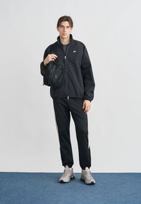 Μαύρο τζάκετ με φερμουάρ και ελαστικά μανίκια, σε συνδυασμό με μαύρο αθλητικό παντελόνι με λευκές ρίγες στο πλάι. Γκρι αθλητικά παπούτσια και μικρή μαύρη τσάντα χιαστί.