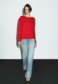 Das Model geht selbstbewusst auf die Kamera zu, trägt einen roten Pullover und helle blaue Jeans, vor einem schlichten weißen Hintergrund und dunklem Boden.
