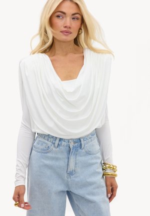 Femme portant un haut blanc drapé et un jean bleu clair taille haute avec des bracelets et une bague en or, regardant sur le côté.