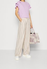 Lila t-shirt med flikiga ärmar, beige randiga vida byxor, vita skor och en lila-mönstrad väska.