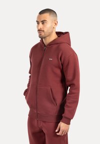 Bordová mikina na zip s prostornou klokaní kapsou, ribbovanými manžetami a spodním lemem, vyrobená z měkké, texturované tkaniny. Klasický střih.
