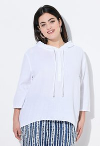 Hoodie bianco con tessuto a trama, chiusura a bottoni e cordini regolabili. Abbinato a un pantalone a righe blu e bianche.