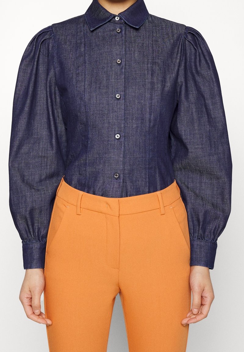 Camicia di jeans navy con colletto e maniche a palloncino, con chiusura a bottoni e leggera lucentezza, abbinata a pantaloni arancioni aderenti.
