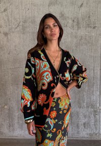 Γυναίκα με μακριά καστανά μαλλιά, φοράει μαύρη ζακέτα και φούστα με ζωηρά πορτοκαλί, μπλε και πράσινα σχέδια παϊέτας, στέκεται μπροστά σε έναν τσιμεντένιο τοίχο.