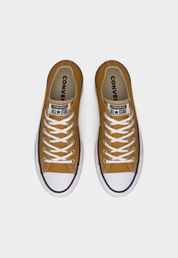 Ζεύγος καφέ χαμηλών παπουτσιών Converse All Star με λευκά κορδόνια και λαστιχένιες μύτες, που φαίνονται από πάνω σε λευκό φόντο.