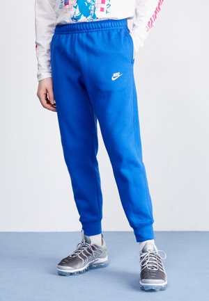 Person trägt leuchtend blaue Nike-Sweatpants, ein weißes langärmeliges Shirt mit Grafik, weiße Socken und graue Sportschuhe und steht auf einem blauen Boden.