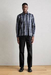 Camisa de botão de manga longa em cinza escuro com padrões verticais abstratos em preto, combinada com calças pretas e sapatos pretos.
