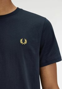 Camiseta de algodón de color azul marino con mangas cortas, que presenta un pequeño bordado de una corona de laurel dorada en el lado izquierdo del pecho.