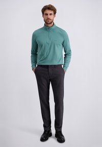 Homme debout les mains dans les poches, portant un pull zippé teal à quart, un pantalon gris foncé et des chaussures habillées noires sur un fond uni.