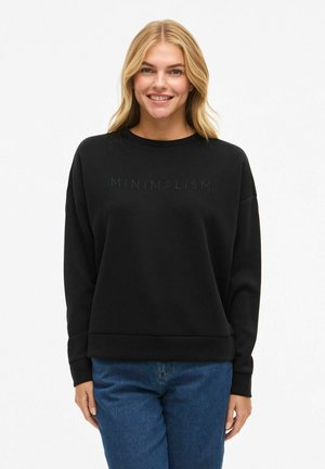 Femme blonde souriante portant un sweat noir avec le texte "MINIMALISME" et un jean bleu, se tenant devant un fond blanc uni.