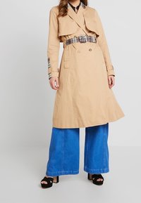 Una donna che indossa un trench beige con cintura sopra jeans a gamba larga blu e tacchi a piattaforma neri aperti, in piedi contro uno sfondo semplice.
