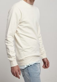 Sudadera crema, cuello redondo, puños y dobladillo acanalados, textura suave, combinada con jeans de color azul claro, vista lateral que muestra el ajuste y el diseño.