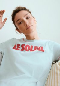 Ljusblå bomulls t-shirt med korta ärmar, med texten "LE SOLEIL" i feta röda bokstäver. Kort hals med en rosa kantdetalj.
