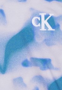 Bawełniany t-shirt z niebiesko-białym abstrakcyjnym wzorem moro. Na lewym górnym rogu znajduje się małe białe logo „cK”. Miękka tekstura.