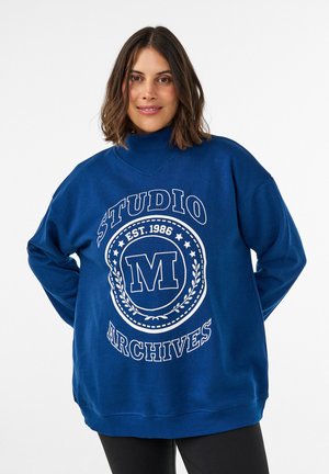 Mujer con cabello castaño hasta los hombros que lleva una sudadera azul con texto y emblema "Studio Archives Est. 1986", de pie con las manos detrás de la espalda.