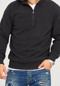 Felpa nera in pile con colletto a mezza zip, polsini e orlo a coste; indossata sopra una camicia bianca, abbinata a jeans blu strappati.