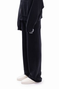Black knitted sweatshirt and matching pants with a relaxed fit. Pants feature a side pocket and small white logo detail. Shoes are light-colored.