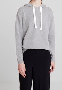 Haut à capuche gris en tissu tricoté doux avec des cordons blancs, un décolleté en V et des poignets côtelés. Associé à un pantalon large noir.