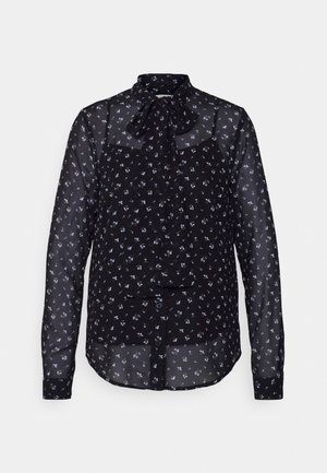 Svart genomskinlig blus med långa ärmar, liten lila blommönster och en knuten rosett vid halslinningen, knäppt framtill.