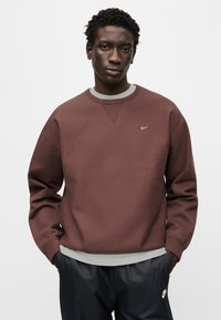 Μπορντό φούτερ με στρογγυλή λαιμόκοψη, ριμπ μανσέτες και γκρι λεπτομέρεια στο κάτω μέρος. Διαθέτει μικρό λευκό λογότυπο Nike στο στήθος.