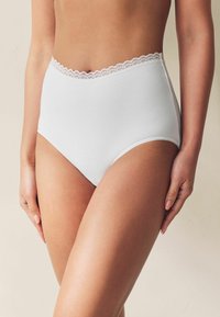 Balti augsta vidukļa kokvilnas biksītes ar viļņotu mežģīņu jostu, gludu tekstūru un bezšuvju sāniem, attēlotas modeļa apakšējā ķermeņa daļā.