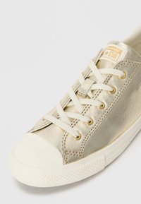 Gouden metallic canvas sneaker met witte rubberen neus en zool, voorzien van platte veters, gouden gaatjes en een klein logo label op de tong.