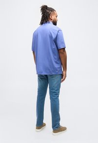 Άνδρας με κοτσιδάκια που φοράει μοβ πόλο μπλούζα, μπλε τζιν και μπεζ αθλητικά παπούτσια, όρθιος με την πλάτη προς τα εμάς σε απλό λευκό φόντο.