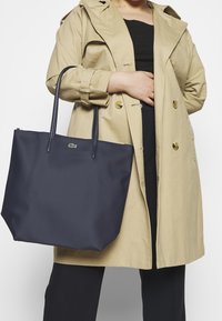 Marinblå tote-väska med en strukturerad yta och två långa handtag. Har en liten grön Lacoste-logga på framsidan.