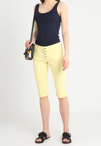 Top sin mangas en color azul marino, pantalones capri amarillos con cinturón y puños enrollados, sandalias negras y un pequeño bolso cruzado negro con detalle de borlas.