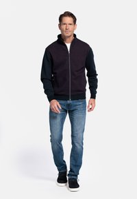 Heren zip-up jas met een donkerpaarse textuur aan de voorkant en zwarte mouwen. Gecombineerd met lichtblauwe aansluitende jeans en zwarte schoenen.