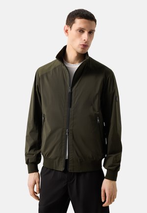 Man die een olijfgroene jas met rits en hoge kraag draagt en een zwarte broek, staand tegen een effen lichte achtergrond.