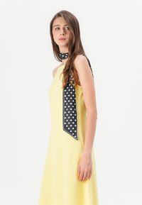 Robe sans manches jaune avec une surface texturée, assortie d'un foulard à pois noirs noué autour du cou, avec une extrémité pointue.