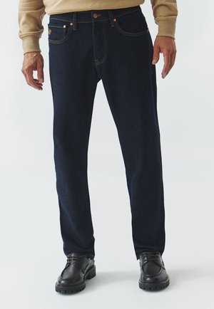 Jeans de mezclilla azul oscuro con detalles de costura, diseño de pierna recta, estilo de cinco bolsillos y cierre de botón. Llevados con zapatos negros.