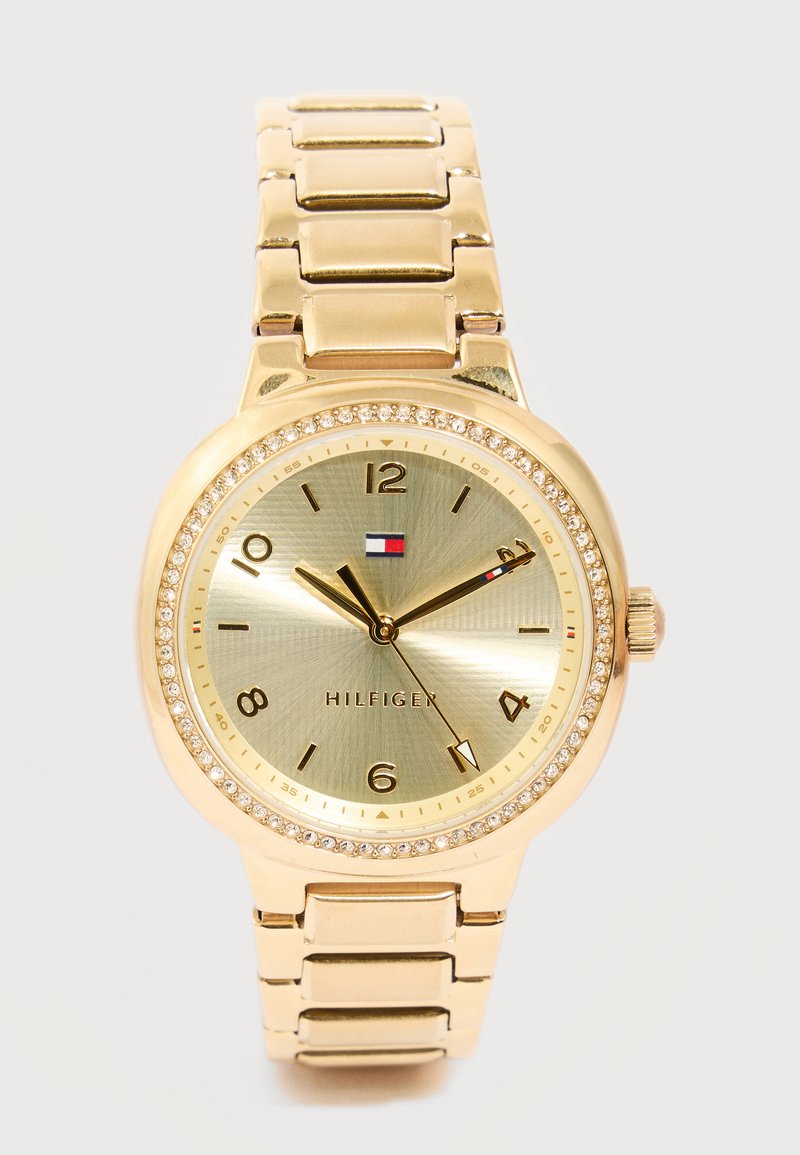 Χρυσό ρολόι χειρός Tommy Hilfiger με στεφάνη από κρύσταλλο, χρυσό καντράν, αριθμητικούς δείκτες ωρών και μπρασελέ από συνδεδεμένο μεταλλικό λουράκι.