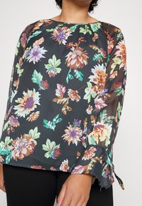 Sort bluse med blomsterprint, der har livlige blomster og blade i grøn, orange, lilla og hvid. Gennemsigtigt stof med rynket halsudskæring og manchetter.
