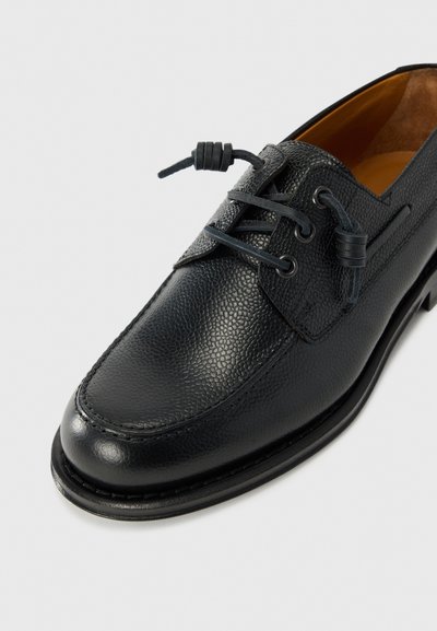 Chaussure en cuir noir avec une surface texturée, bout arrondi et laçage décoratif avec des accents noués. Semelle robuste avec des détails de couture.