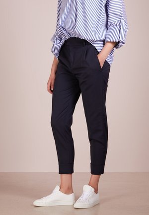 Osoba ubrana w granatowe zwężane spodnie z podwiniętymi nogawkami, niebiesko-białą paskowaną bluzkę oraz białe trampki, stojąca z jedną ręką w kieszeni.