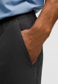 Pravá ruka zasunutá vrecku čiernych nohavíc s elastickým pásom, oblečená v svetlomodrej košeli, na jednoduchom svetlom pozadí.