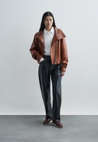 Smeđa kožna jakna sa širokim ovratnikom, uparena s crnim hlačama širokih nogavica i bordo cipelama. Model stoji na minimalističkoj pozadini.
