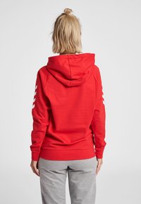 Felpa rossa con cappuccio e vestibilità morbida, caratterizzata da strisce bianche sulle maniche e una texture morbida. Abbinata a pantaloni grigi, che mostrano la vista posteriore.