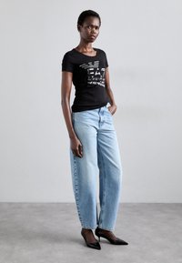 Zwarte korte mouwen t-shirt met reflecterend logo, gecombineerd met lichtgewassen rechte jeans en zwarte puntige hakken. Gladde texturen.