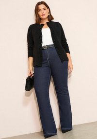 Frau trägt schwarzen, geknöpften Cardigan, weißes Oberteil, dunkelblaue High-Waist-Jeans mit Gürtel, hält schwarze Handtasche, steht vor beiger Wand.