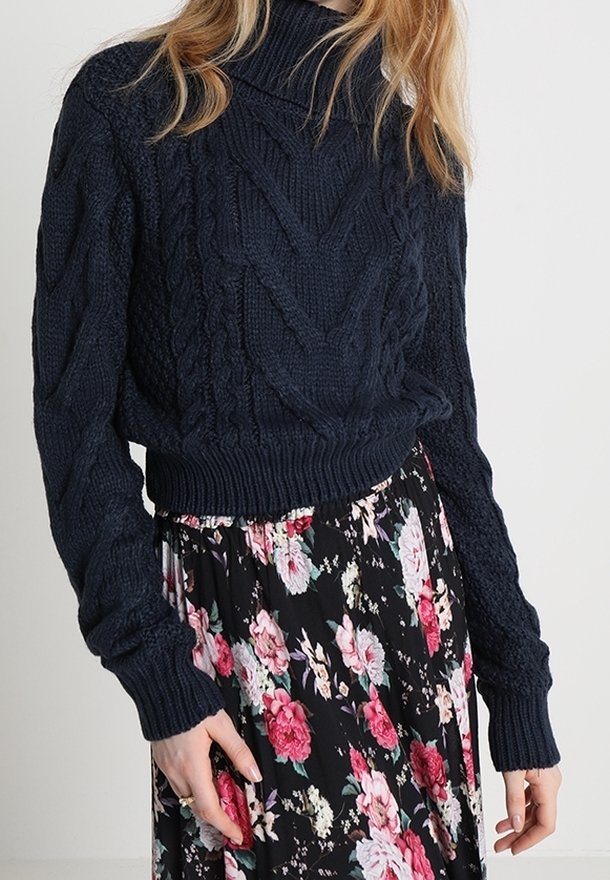 Femme portant un pull à col roulé en maille torsadée bleu marine foncé et une jupe noire à fleurs roses et blanches.