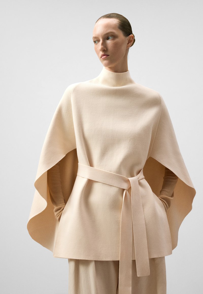 Beige cape-achtige top gemaakt van zachte stof, met een hoge hals, brede mouwen en een bijpassende riem die om de taille is geknoopt.
