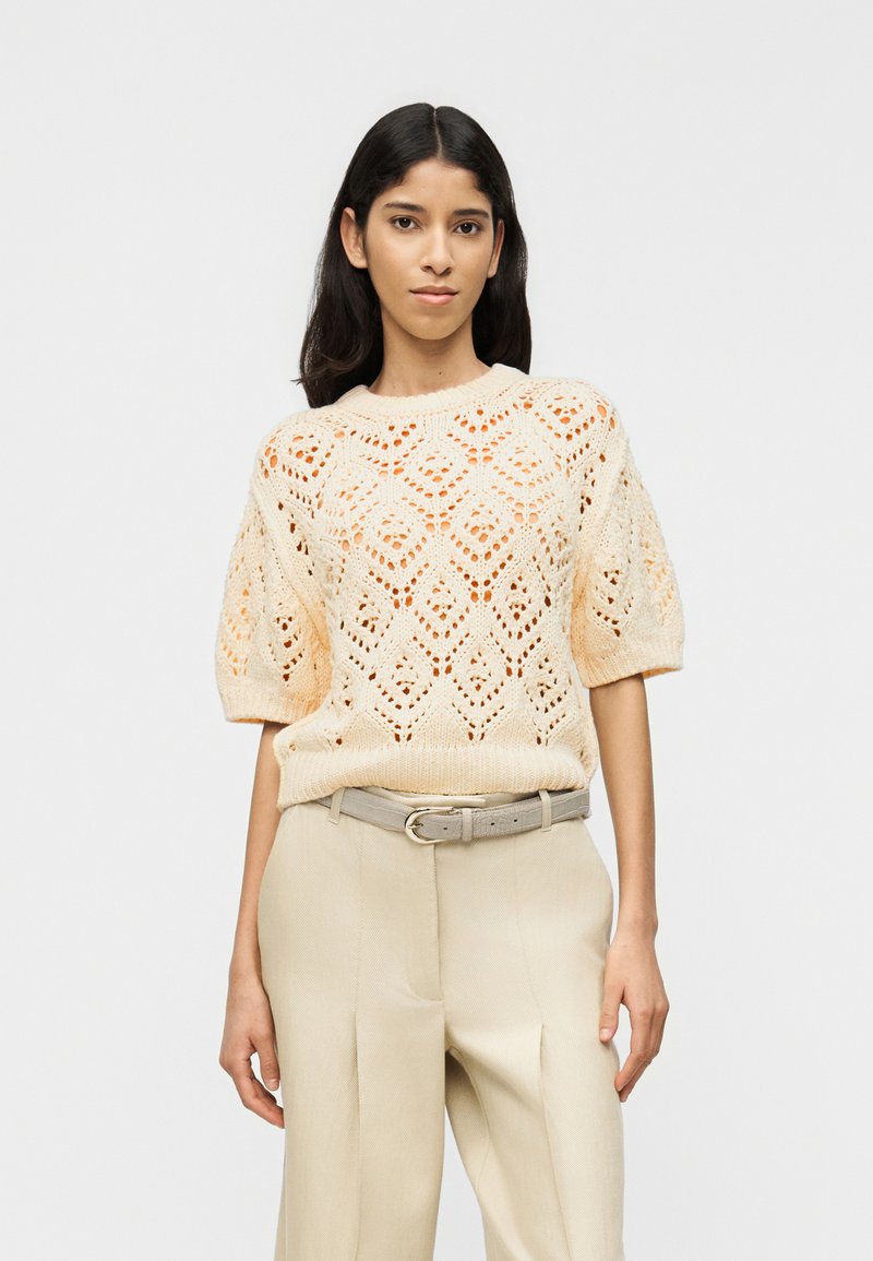 Jeune femme debout portant un pull court à manches courtes en maille crème et un pantalon taille haute beige avec une ceinture assortie, sur un fond uni.