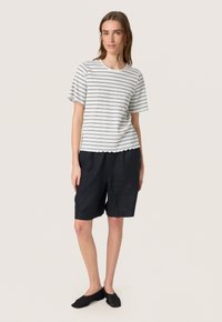 Top de manga corta a rayas con dobladillo ondulado en blanco y negro, combinado con pantalones cortos de lino azul marino oscuro. Unos zapatos negros sin cordones completan el look.