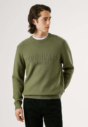 Suéter verde de punto con puños y dobladillo acanalados. Presenta un logotipo texturizado en un tono más claro. Combinado con pantalones negros. Diseño clásico de cuello redondo.